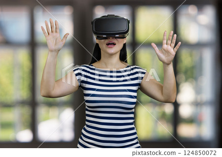 Amazed woman in vr headset with her hands up. Blurred checkered windows background. 124850072