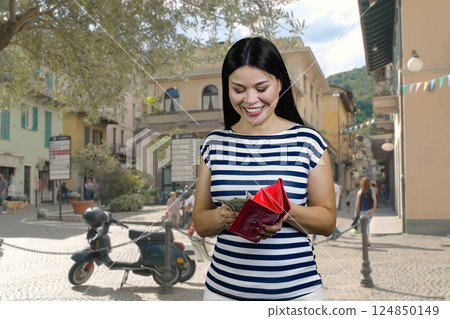 Smiling cheerful asian woman is getting money from red wallet. Standing outdoors on the street. Smiling cheerful asian woman is getting money from red wallet. Standing outdoors on the street. 124850149