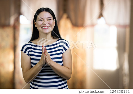 Happy cheerful asian woman with praying hands gesture. Standing indoors on windows backgound. 124850151