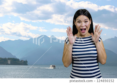 Asian brunette woman is screaming in fear standing outdoors. Sea and mountain scape background. Asian brunette woman is screaming in fear standing outdoors. Sea and mountain scape background. 124850152