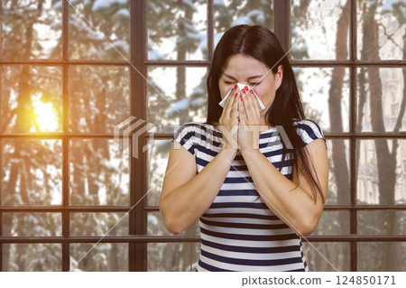 Asian woman with runny nose standing indoors. Sneezing brunette on checkered windows background with winter evening landscape. 124850171