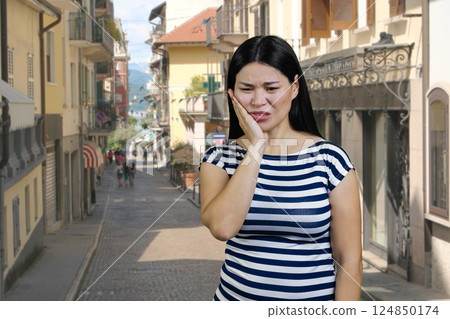 Young asian woman suffering from toothache outdoors. Italian european street background. 124850174