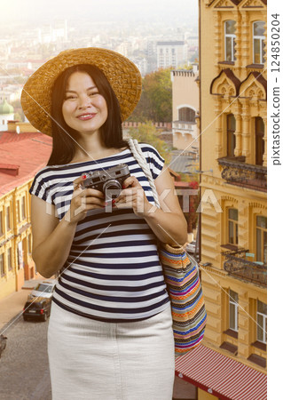 Vertical shot of asian woman tourist in a european street. Cheerful korean girl in straw hat is holding a photo camera. Vertical shot of asian woman tourist in a european street. Cheerful korean girl in straw hat is holding a photo camera. 124850204