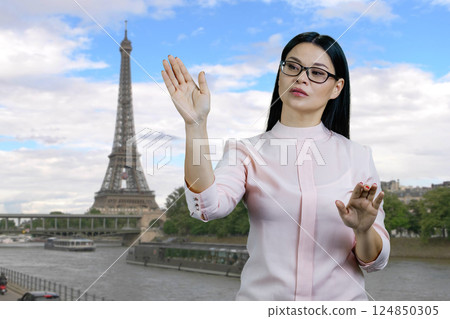Young asian business woman swipes invisible virtual screen standing outdoors. France Eiffel Tower background. 124850305