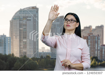 Young asian woman is touching an invisible virtual with her hand. Urban cityscape background. Young asian woman is touching an invisible virtual with her hand. Urban cityscape background. 124850307
