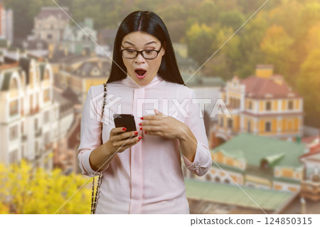 Shocked and surprised asian young woman is looking at her smartphone screen. Businesswoman standing outdoors. European town background. 124850315