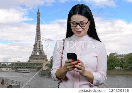 Portrait of a young asian woman texting something on smartphone standing outdoors. Paris Eiffel Tower background. Portrait of a young asian woman texting something on smartphone standing outdoors. Paris Eiffel Tower background. 124850326