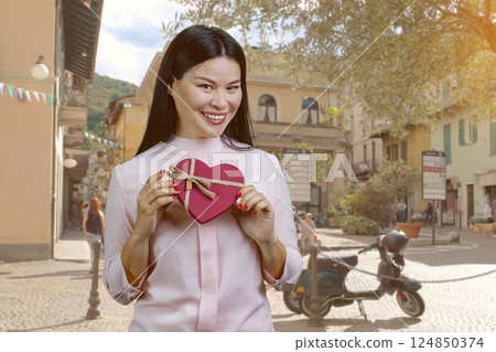 Portrait of smiling asian woman holding a red heart shaped gift box. European street urban scape background. 124850374