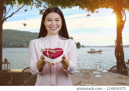 Smiling cheerful asian woman showing heart shape gift box. Outdoor resort landscape background. 124850375