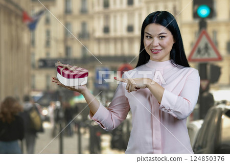 Young asian woman with heart shape gift box standing outdoors. Strees scape background. Young asian woman with heart shape gift box standing outdoors. Strees scape background. 124850376