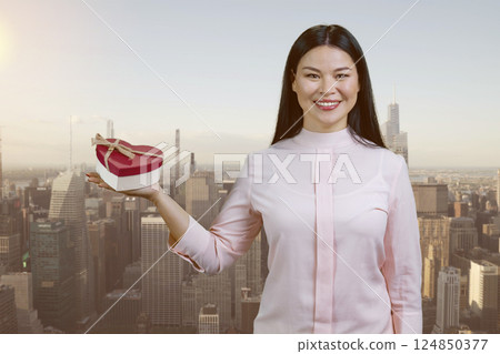 Young asian woman is showing a heart shaped gift box. Urban cityscape backgorund. Young asian woman is showing a heart shaped gift box. Urban cityscape backgorund. 124850377