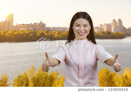 Portrait of young smiling asian woman giving two thumbs up. Outdoor city river background. 124850418