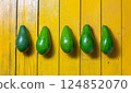 Fresh green avocados lined up on a vibrant yellow wooden surface during daylight 124852070
