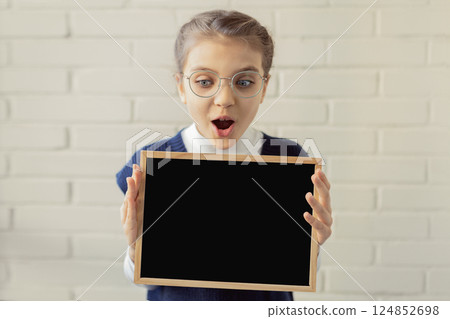 Surprised happy 8 year old girl wearing glasses and business clothes holds black mockup chalk board on white brick wall background, copy space Surprised happy 8 year old girl wearing glasses and business clothes holds black mockup chalk board on white brick wall background, copy space 124852698