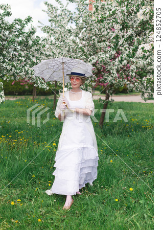 Natural adult mature woman of 60 years old in 18th century style white canotier hat, retro white dress, gloves, glasses and umbrella in blossoming white and pink apple orchard on summer day Natural adult mature woman of 60 years old in 18th century style white canotier hat, retro white dress, gloves, glasses and umbrella in blossoming white and pink apple orchard on summer day 124852705