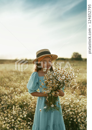 Portrait of natural girl in blue linen dress and canotier straw hat stands and holds bouquet of flowers in daisy field, vintage photo 124852795