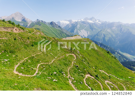 Scenic aerial panoramic view curvy winding serpentine dirt path road alpine mountain peak with green wild grassland meadow and rocky horizon sky landscape. Hill nature scenery hiking route sunny day Scenic aerial panoramic view curvy winding serpentine dirt path road alpine mountain peak with green wild grassland meadow and rocky horizon sky landscape. Hill nature scenery hiking route sunny day 124852840