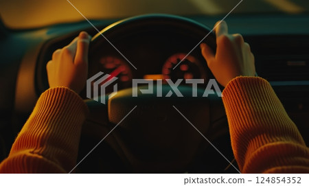 Close up of woman's hands gripping the steering wheel while driving through the night, with the dashboard softly illuminated by an inviting orange glow, creating a warm ambiance 124854352
