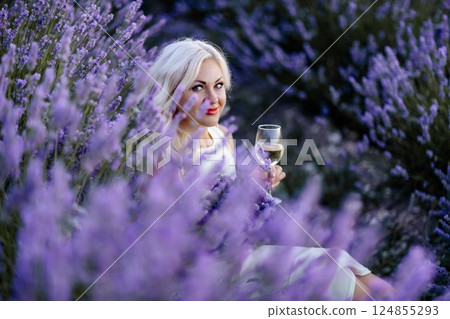 Blonde lavender field holds a glass of white wine in her hands. Happy woman in white dress enjoys lavender field picnic holding a large bouquet of lavender in her hands . Illustrating woman's picnic 124855293