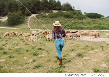 A woman wearing a hat and jeans walks through a field of sheep 124855420
