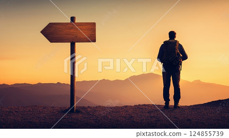 Male hiker with backpack standing on mountain top at sunset looking at mountain range with blank wooden signpost indicating choice of direction 124855739