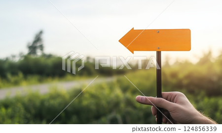 Hand holding blank orange directional arrow sign pointing left with blurred background of road and nature at sunset, symbolizing choice, decision making, way, and direction 124856339