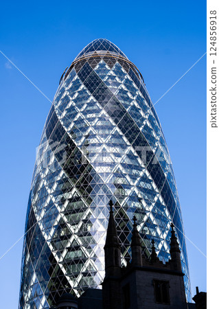 Gherkin Building rises prominently against a clear blue sky in London, showcasing its unique, curved glass architecture. 124856918