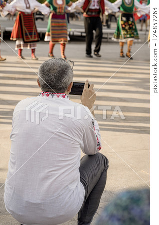 Man squats with smartphone to capture traditional dancers performing in ethnic costumes. People in traditional folk costumes holding hands while dancing in cultural Bulgarian festival 124857263