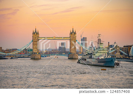 The iconic Tower Bridge stands majestically over the Thames River as HMS Belfast rests nearby. The evening sky glow envelops London, creating a picturesque scene. 124857408