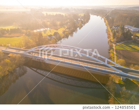 An elegant bridge arches over the tranquil Labe River in Litol, Lysa nad Labem, Czechia. The soft morning light enhances the lush green landscape surrounding the waterway. 124858501