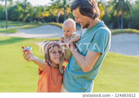 Father with his two sons in the park, the older boy wearing a pilot hat and holding a toy airplane while the baby watches. Childhood, bonding, and family adventure concept Father with his two sons in the park, the older boy wearing a pilot hat and holding a toy airplane while the baby watches. Childhood, bonding, and family adventure concept 124863812
