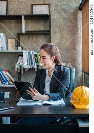 Professional woman in office setting using a tablet, with a safety helmet on the desk, symbolizing industry and technology. Professional woman in office setting using a tablet, with a safety helmet on the desk, symbolizing industry and technology. 124865568