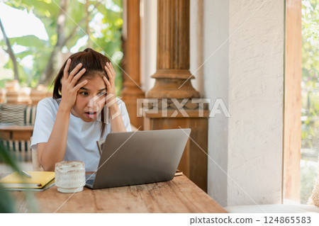 A young woman looks stressed while working on her laptop at home, surrounded by coffee and a notebook on a wooden table. 124865583