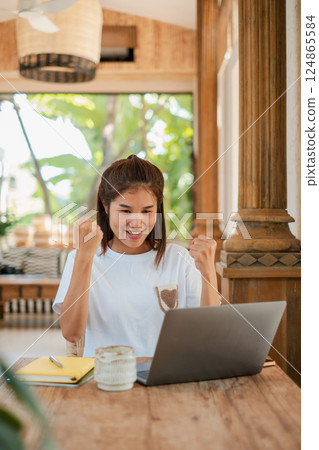 A joyful young woman celebrates a success while using her laptop in a bright, cozy home office setting. 124865584