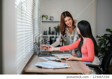 Two women working together in a bright office, discussing ideas with laptops and documents on a wooden desk. 124865599