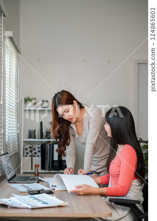 Two women working together in a bright office, reviewing documents and using a laptop, showcasing teamwork and productivity. 124865601
