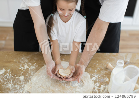 Caucasian family including adult and child preparing cookies in kitchen holding heart shape cutout together. Image highlights bonding, togetherness, and fun in culinary activities. Caucasian family including adult and child preparing cookies in kitchen holding heart shape cutout together. Image highlights bonding, togetherness, and fun in culinary activities. 124866799