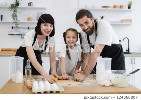 Caucasian family, parents and daughter, prepare cookies by cutting dough with molds in kitchen. Fun, teamwork, quality time, homemade baking atmosphere. Caucasian family, parents and daughter, prepare cookies by cutting dough with molds in kitchen. Fun, teamwork, quality time, homemade baking atmosphere. 124866807