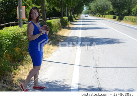 Young woman in blue sling dress with baby standing on the side of the road 124868083