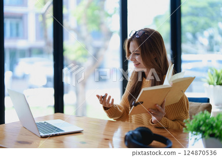 Smiling young woman engaged in a joyful online e-learning video call, gestu while holding a book with headphones on a wooden table nearby 124870536