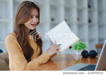 Smiling young woman engaged in a joyful online e-learning video call, gestu while holding a book with headphones on a wooden table nearby Smiling young woman engaged in a joyful online e-learning video call, gestu while holding a book with headphones on a wooden table nearby 124870541