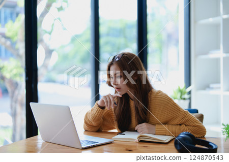 Smiling young woman engaged in a joyful online e-learning video call, gestu while holding a book with headphones on a wooden table nearby Smiling young woman engaged in a joyful online e-learning video call, gestu while holding a book with headphones on a wooden table nearby 124870543