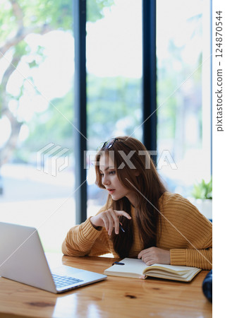 Smiling young woman engaged in a joyful online e-learning video call, gestu while holding a book with headphones on a wooden table nearby Smiling young woman engaged in a joyful online e-learning video call, gestu while holding a book with headphones on a wooden table nearby 124870544