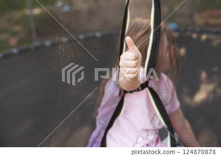 Happy excited child girl sitting on trampoline outdoor showing thumb up sign 124870878