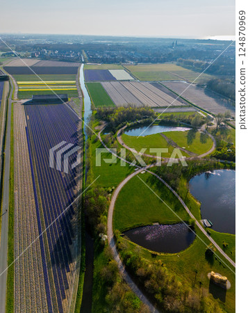 Aerial view of scenic farmland near the coast with colorful flower fields, dense forests, and circular ponds in a landscaped park. The North Sea is visible in the distance under a clear sky. Aerial view of scenic farmland near the coast with colorful flower fields, dense forests, and circular ponds in a landscaped park. The North Sea is visible in the distance under a clear sky. 124870969