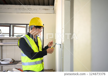 Electrician wearing yellow hard hat working on wall wiring at a construction site 124873376