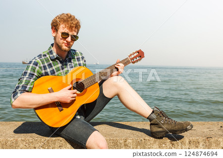 Young man hipster playing guitar by sea ocean. Young man hipster playing guitar by sea ocean. 124874694