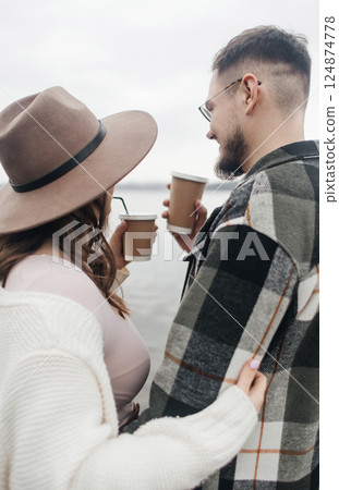 Young couple on a date. A woman in the hat and a man with glasses stand looking at the water, hugging and drinking coffee from disposable cups. 124874778