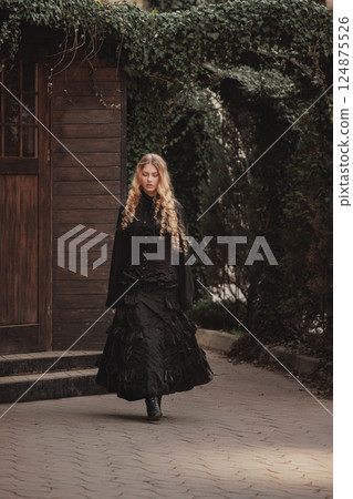 Young woman with golden curls in black lace gown walks slowly under ivy arch beside wooden house. Suitable for dark fairytale or novel cover. 124875526