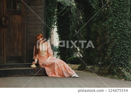 Sitting near wooden door, head bowed, she clasps an open book, vines curling around her like nature's embrace. Sitting near wooden door, head bowed, she clasps an open book, vines curling around her like nature's embrace. 124875554